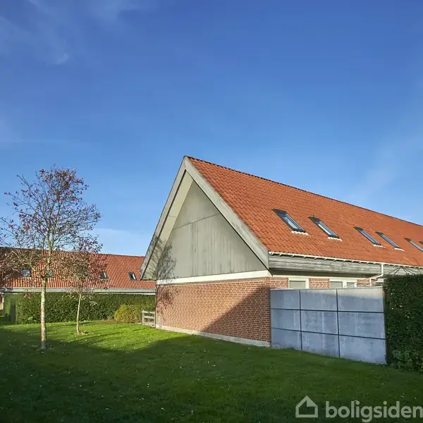 Hus med rødt tegltag står stille i et grønt boligområde med græsplæne og træer. Klar blå himmel dominerer baggrunden.