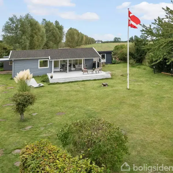 Et gråblåt sommerhus står i en rummelig have med en hvid terrasse og havemøbler. En flagstang med et dannebrog vajer i vinden, omgivet af træer og buske.