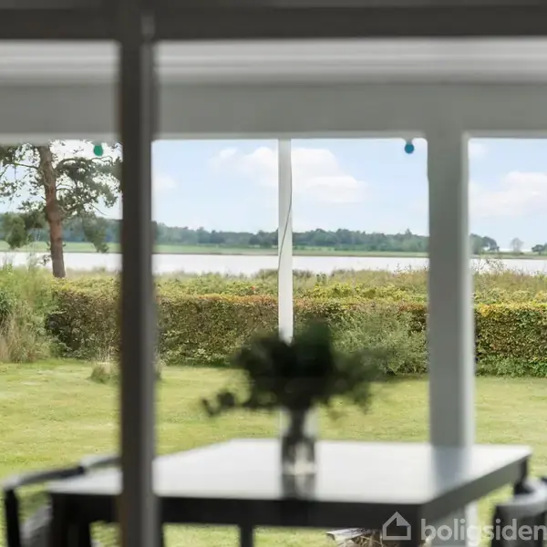 Bord står stille foran store vinduer; blomsterudsmykning i forgrunden. Udsigt til græsplæne, smukke buske og en sø i baggrunden under en skyfri himmel.