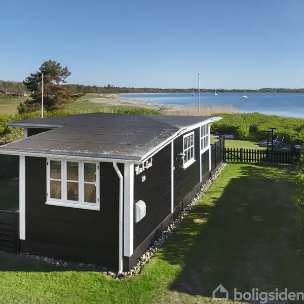 Sortmalet sommerhus med hvide vinduesrammer står stille ved kysten. Omgivet af grønne buske med udsigt over en rolig blå sø eller hav.