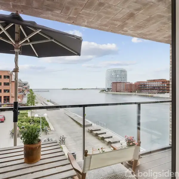 En balkon med planter og parasol udsigt over en moderne byvandkanal med bygninger på begge sider. En stor glasfacade viser det åbne landskab med blå himmel og skyer.
