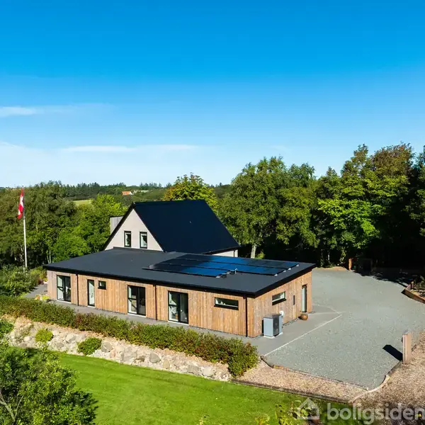 Et moderne hus med fladt tag og solpaneler står i en rummelig, grusbelagt gårdsplads omgivet af frodig skov og en velplejet græsplæne. En dansk flagstang står i nærheden.