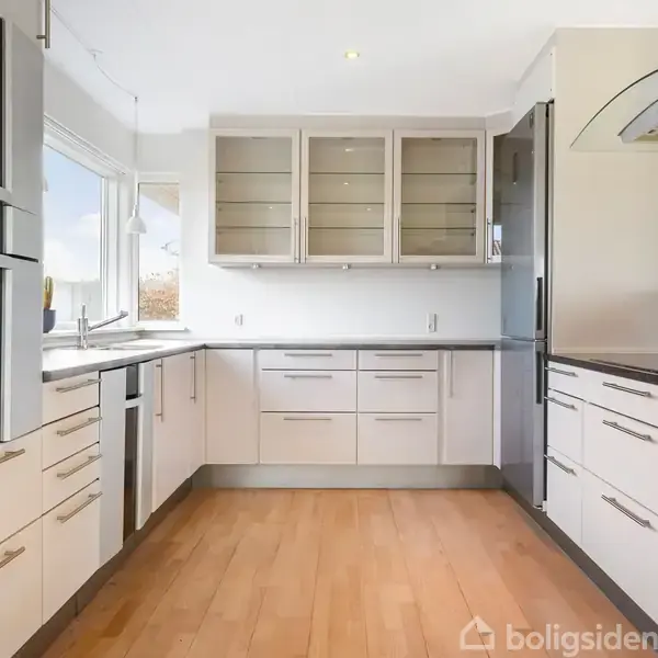 Et moderne køkken med hvide skabe og stålgreb. Køkkenet har en sort bordplade, indbygget ovn og en stor fryser. Rummets lyse trægulv og naturlige lys skaber en rummelig atmosfære.