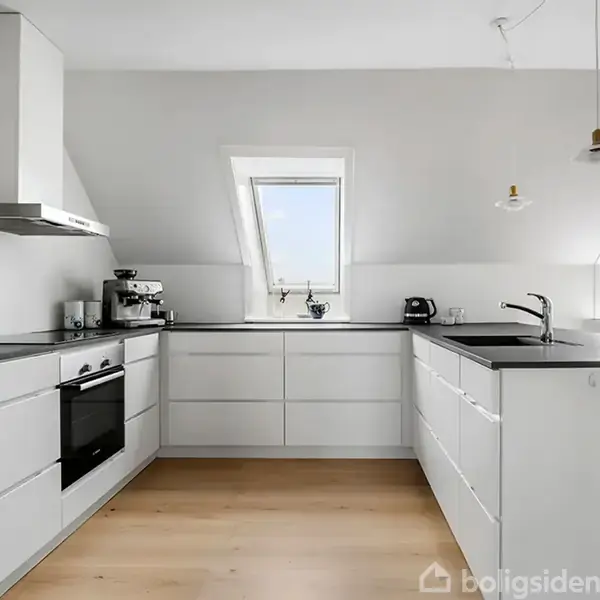 Et moderne, hvidt køkken med indbyggede skabe og apparater, herunder en komfur og emhætte. To ovenlysvinduer slipper lys ind, mens en plante og frugtskål pynter bordpladen.