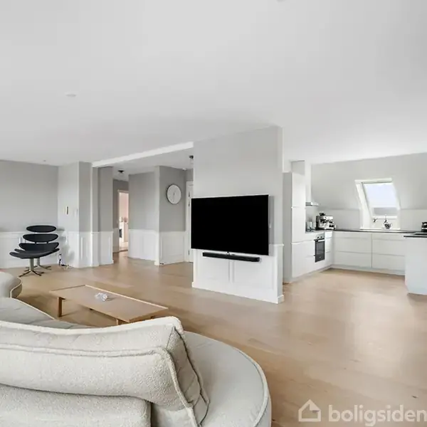 Stue med sofa og tv, moderne køkken i hjørnet, lyst trægulv, og en kamin til venstre. En plante står ved et vindue. Ingen tekst fundet.