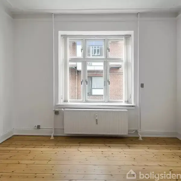 Et tomt rum med et trægulv, en radiator under et vindue, og en hvid væg. Udenfor ses en murstensbygning. Ingen tekst i rummet.