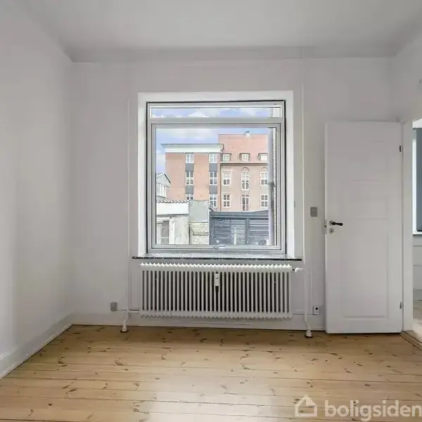Et lyst værelse med trægulv har et vindue, der viser udsigt til en bygning udenfor. Der er en radiator under vinduet og en åben dør til højre.