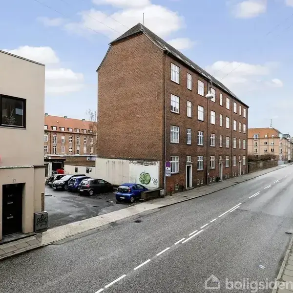 Et vejforløb i en by med en rød murstensbygning til højre. En mindre beige bygning ligger til venstre. Biler er parkeret ved siden af bygningerne. Skyet himmel.