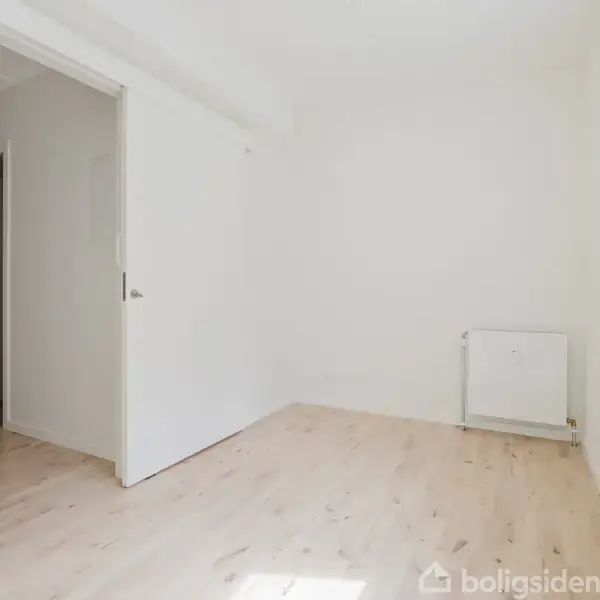 Et tomt, lyst rum med trægulv. En åben dør fører ind til en anden del af boligen. En radiator er monteret på væggen. Ingen tekst er til stede.