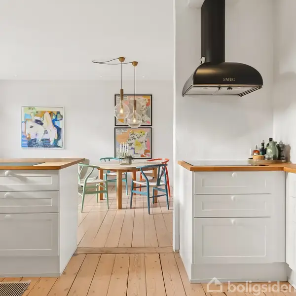 Hvidt køkken med træbordplader og SMEG-emhætte. Køkkenet fører til en spisestue med et rundt træbord og moderne kunst på væggene. Rummene er lyse og minimalistisk indrettet.