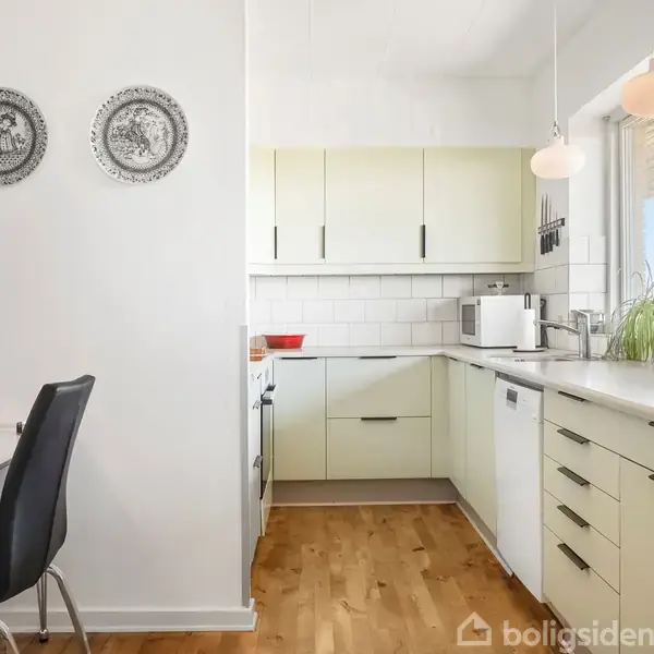 Et lyst køkken med lyse skabe, trægulv, og en hvid bordplade. Der er også en spiseplads med sorte stole og en pendellampe. Vindue til højre og dekorative tallerkener på væggen.