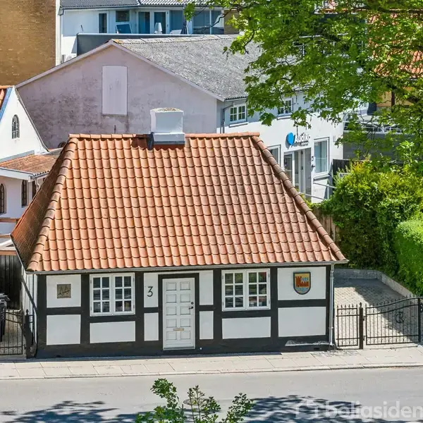 Et bindingsværkshus med rødt tegltag står stille omgivet af grønne buske og træer. Huset har en hvid dør med tallet "3" og et byvåben på væggen.