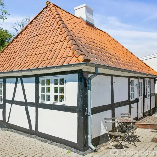 Et bindingsværkshus med rødt tegltag står stille på en brostensbelagt gårdsplads. Ved siden af huset er der et lille bord med to stole, omgivet af et grønt skovområde.