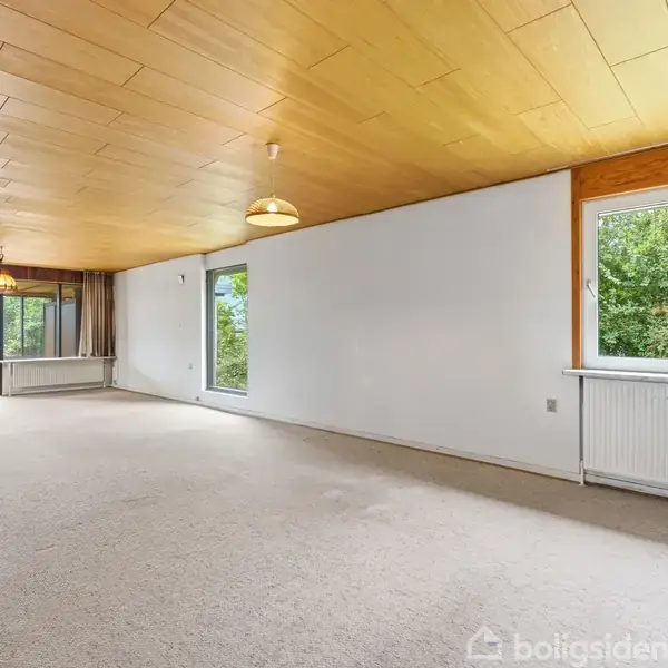 Tomt værelse med beige tæpper, træloft og flere vinduer, der slipper lys ind. Rummet indeholder radiatorer under vinduerne og pendellamper. Vinduerne viser udsigt til grøn beplantning udenfor.