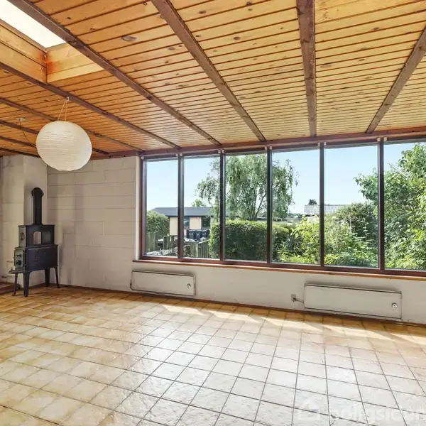 Et rum med en brændeovn i hjørnet, omgivet af store vinduer, der giver udsigt til træer og en blå himmel. Et loft af træ understøtter en stor papirlampe.