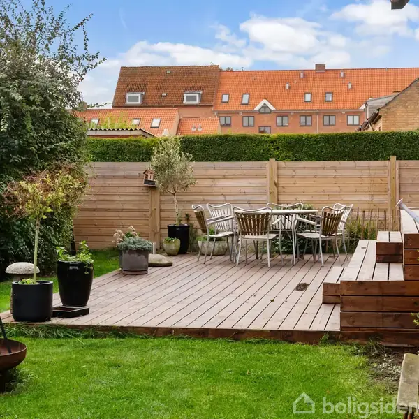 Terrasse i træ står midt i en græsplæne, omkranset af planter og et hegn. Rundt havebord med stole står på terrassen. Baggrunden viser røde hustage under en blå himmel.