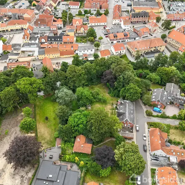 Bebyggelse med røde tage omgivet af frodige træer og grønne områder. En vej snor sig gennem området med flere huse og biler parkeret. Tæt byområde ses i baggrunden.