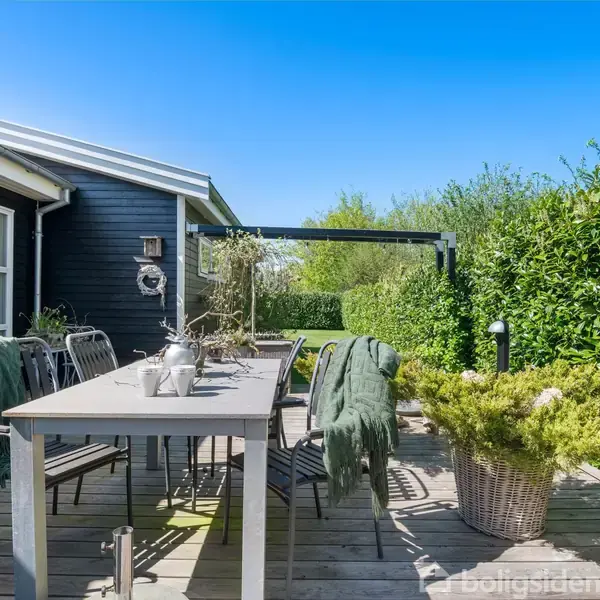 Terrassemøbler står på en træterrasse ved siden af et hus. Flere stole omgiver et bord med dekorationer. En hæk omkranser området under en klar blå himmel.