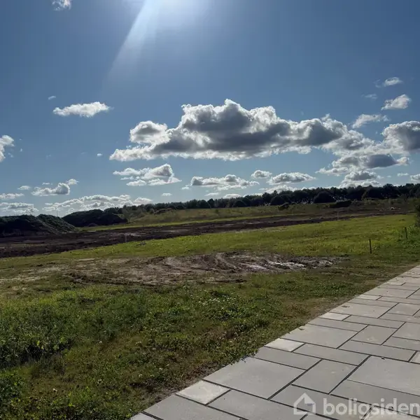 Græsareal ligger uberørt under en blå himmel med spredte skyer. I forgrunden er en flisebelagt sti, mens skov kan ses i horisonten.