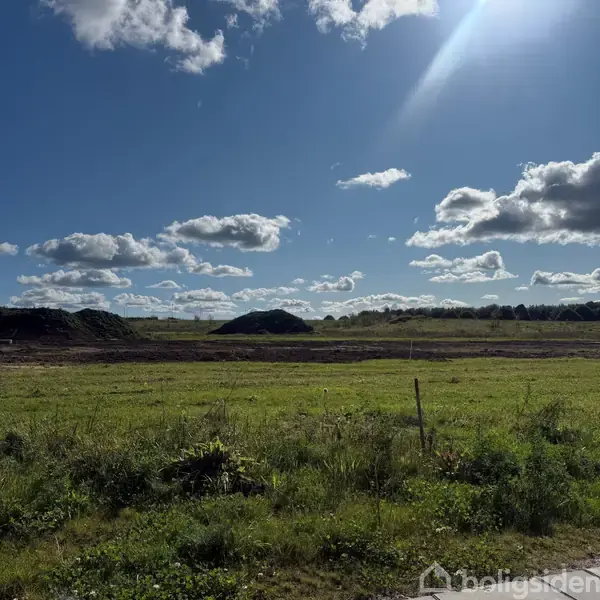 Græsareal strækker sig ud under en blå himmel med spredte, hvide skyer. I baggrunden er lave jordvolde. Det er åbent landskab med skovlinje i horisonten.