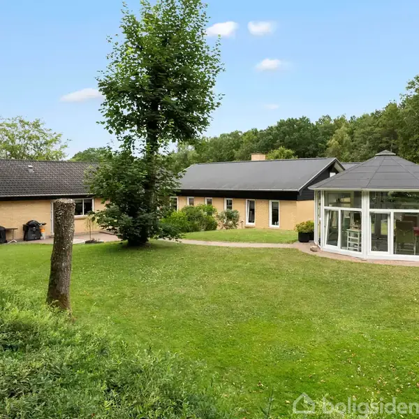 Et gult enfamiliehus med sort tag står i en frodig have med græsplæne. Ved siden af huset er en rund pavillon med glasvægge. Omkring er der træer og buske.