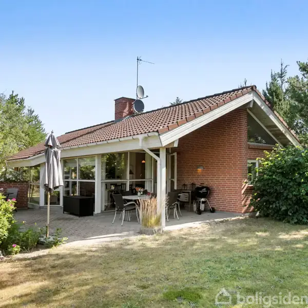 Rødt murstenshus med saddeltag står i en grøn have. To terrassesæt med parasol er placeret på flisebelægning foran huset, der omkranses af træer og buske.
