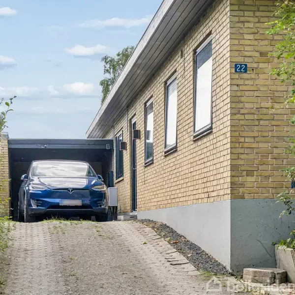 En blå bil holder delvis i en carport ved siden af et gult murstenshus. En indkørsel omgivet af grønne hække leder op til huset. Husnummeret "22" er synligt.
