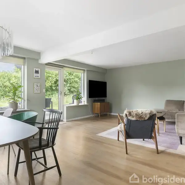 Stue med lysindfald fra store vinduer, grønne planter på vindueskarmen, rundt spisebord med stole, sofa og fladskærms-TV på væggen. Udsigt til grøn have gennem vinduerne.