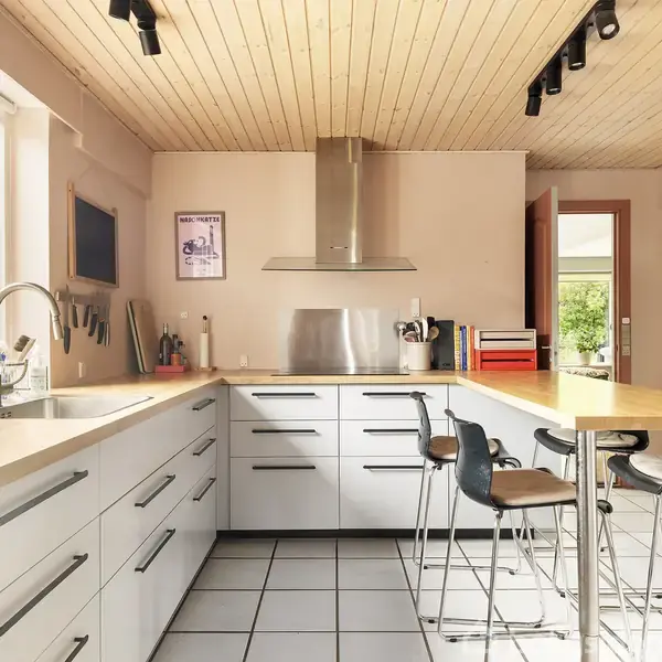 Køkken med lyse hvide skabe og stålgreb. To høje stole ved en træplade. Stor vindue med udsigt til have. Loft af lyst træ med sorte lamper. Retro plakat på væggen.