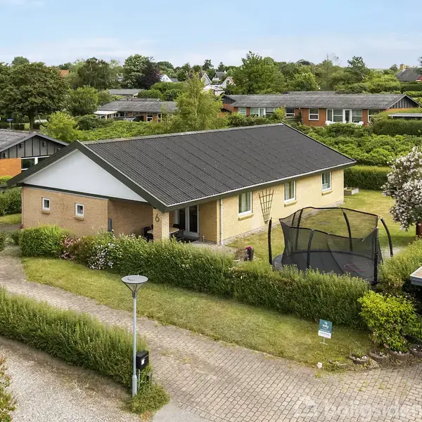 Et gulstenshus med sort tag står stille i en velplejet have, hvor en trampolin er synlig foran. Omkring huset ses en række buske og træer samt nærliggende huse i en villavej.