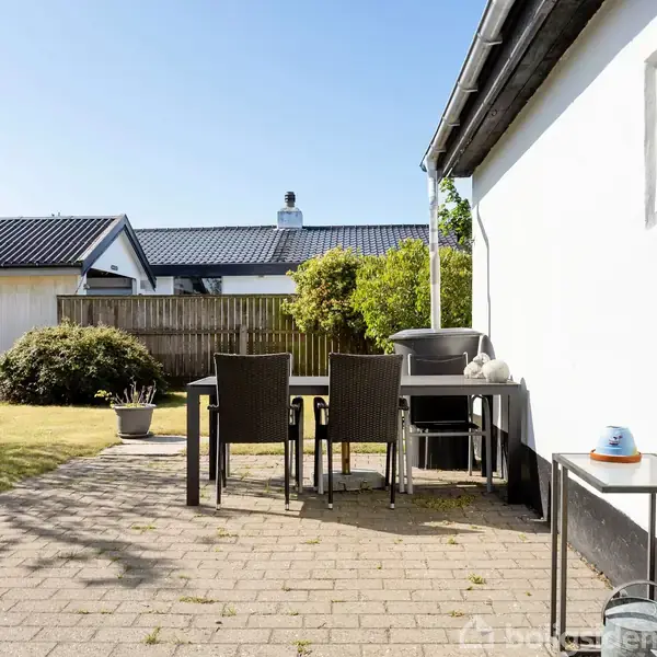 En udendørs terrasse har bord og fire stole foran et hvidt hus. Der står en grill ved siden af bordet, og der er planter og buske i baggrunden.