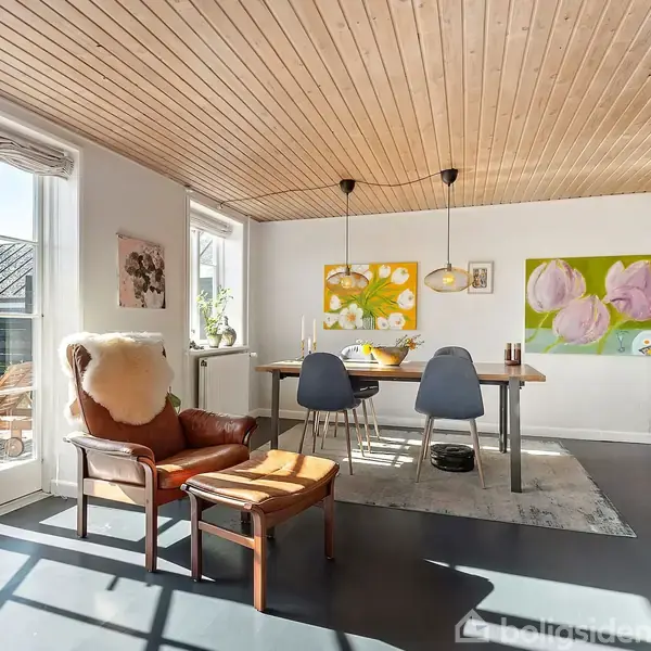 En stue med en brun lænestol og skammel foran store vinduer. Et spisebord med stole. På væggen hænger malerier. Brændeovn opvarmer rummet. Sollys strømmer ind.