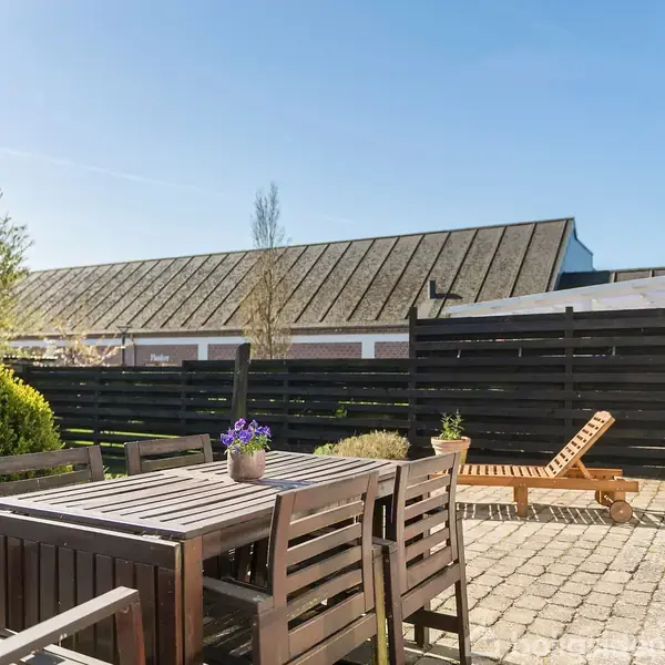 Træbord med stole står på en flisebelagt terrasse ved siden af liggestol. Solbeskinnet have omgivet af buske, træer og sort træhegn. En bygning ses i baggrunden.