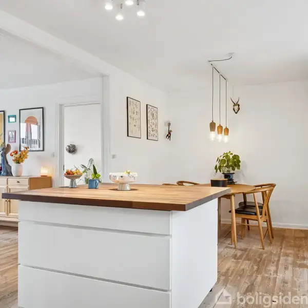 Et hvidt køkkenbord med træoverflade står centralt med frugtskål og påskeliljer. Der er lyse vægge med billeder, træmøbler, og en grøn plante i hjørnet.