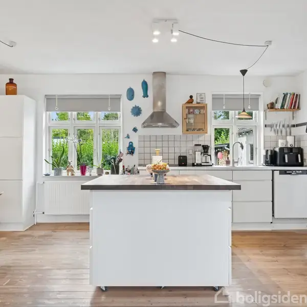 Et lyst køkken med en stor ø i midten. Der er moderne hvide skabe, en ovn og mikrobølgeovn til højre, vinduer med planter, og trægulve. Ingen tekst til stede.