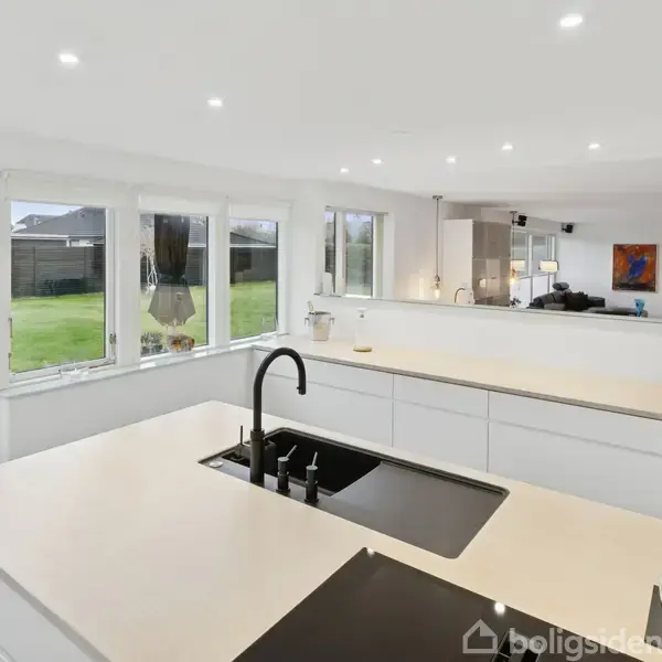 Et moderne køkken med sort håndvask og induktionskogeplade. Køkkenet har store vinduer med udsigt til en grøn have, og i baggrunden ses en åben stue med sofaer og kunst på væggen.