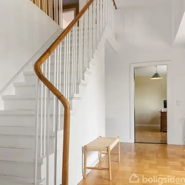Hvid trappe med trægelænder stiger opad. I baggrunden ses en døråbning, der fører til et rum med træmøbler og en loftslampe. En bænk står i gangen.