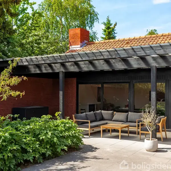 Terrasse med sort sofaarrangement og træbænk står under pergola. Rød murstenshus omkranser området. Grønne buske og træer omkranser terrassen og skaber en hyggelig baggårdsatmosfære.