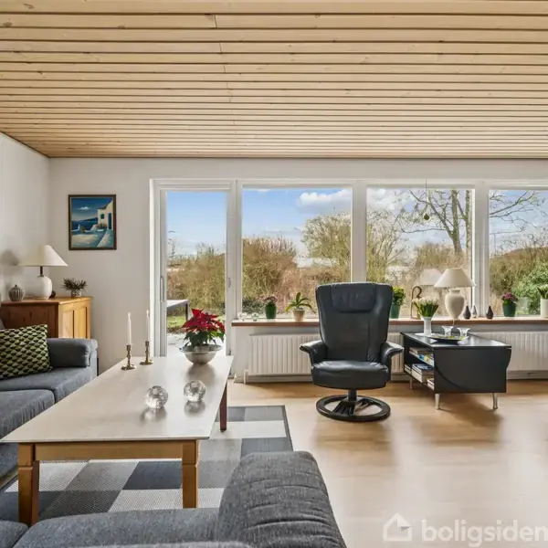 Stue med grå sofa og lænestole omkring et sofabord, udsigt gennem store vinduer til en have. Vægpynt og lamper skaber en hyggelig atmosfære.