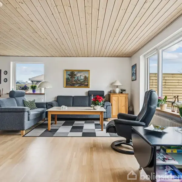 Stue med grå sofa og lænestol på sort-hvidternet tæppe, flankeret af træmøbler. Store vinduer giver udsigt til have. Plakater og malerier pryder hvide vægge, og grønne planter står i vindueskarmen.