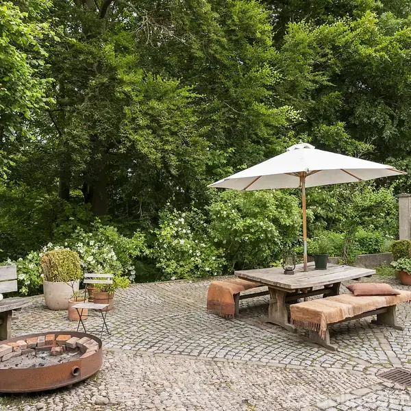 Borde-bænkesæt under parasol står på brostensbelagt terrasse. Området inkluderer ildsted og potteplanter. Omgivet af frodig grøn bevoksning og en hvid husvæg med bruser.