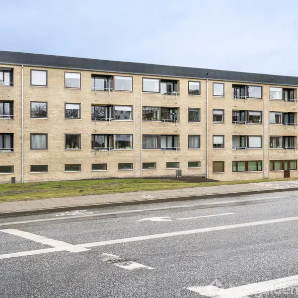 En fleretagers boligbygning står i en byområde, bygget i gule mursten med store vinduer. Foran løber en bred vej med flere markerede kørebaner og cykelsti til venstre.