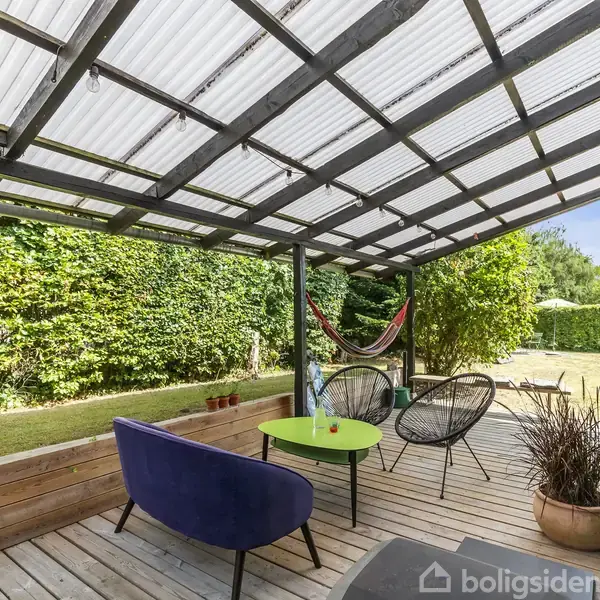 Terrasse med en overdækning i gennemsigtigt materiale. To stole og en blå sofa står omkring et grønt bord. En hængekøje hænger i baggrunden. Haven er omgivet af grønne hække.