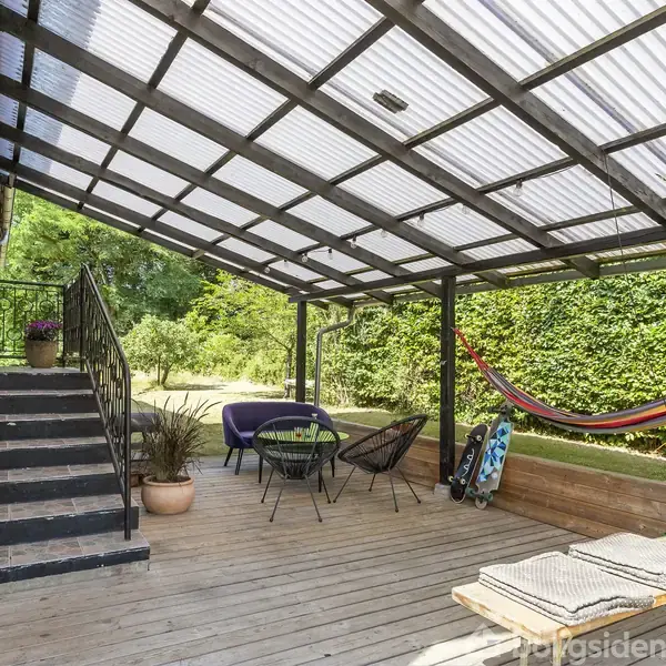 Terrasse med pergola af metal og gennemsigtigt tag. Der er trapper, potteplanter, borde, stole og en hængekøje. Omgivet af en grøn have og hække.