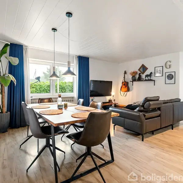 Spisebord med stole er i centrum, omgivet af en stue med sofa, tv og guitar på væggen. Store vinduer med blå gardiner giver lys til rummet.