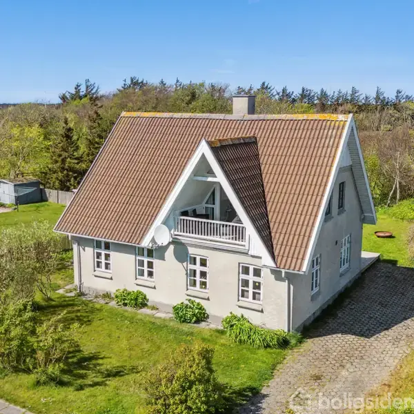 Et gråmalet hus med rødt tegltag står på en grøn græsplæne. Huset har en altan, og omgivende træer ses i baggrunden.
