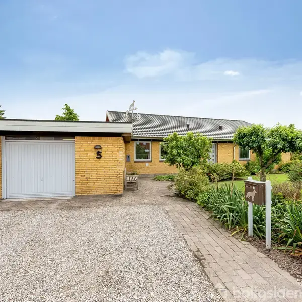 Graveret stenhus står stille i en velplejet have. Grusindkørsel fører til en garage med en dør. "5" ses ved garagen. Blå himmel i baggrunden.