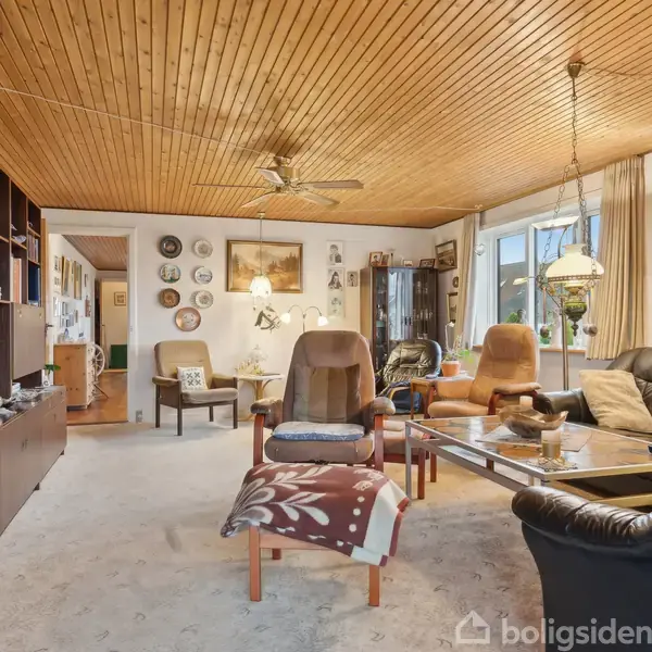 Stue med sofaer og lænestole, der vender mod sofabord i glas. Træloft med ventilator. Reol med bøger og pyntegenstande til venstre. Vinduer og malerier på væggen.