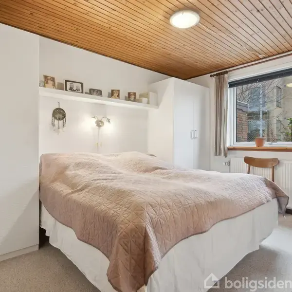 En seng med beige sengetæppe står i et lyst soveværelse med træloft. Væggene har indbyggede skabe, og et vindue i baggrunden giver udsigt til murstensbygning udenfor.