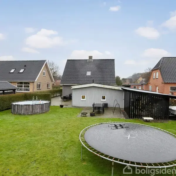 Trampolin og swimmingpool står stille i en grøn have med et lille skur og haveborde. Omkranset af hegn og tre huse med klassiske tagformer under en lyseblå himmel.